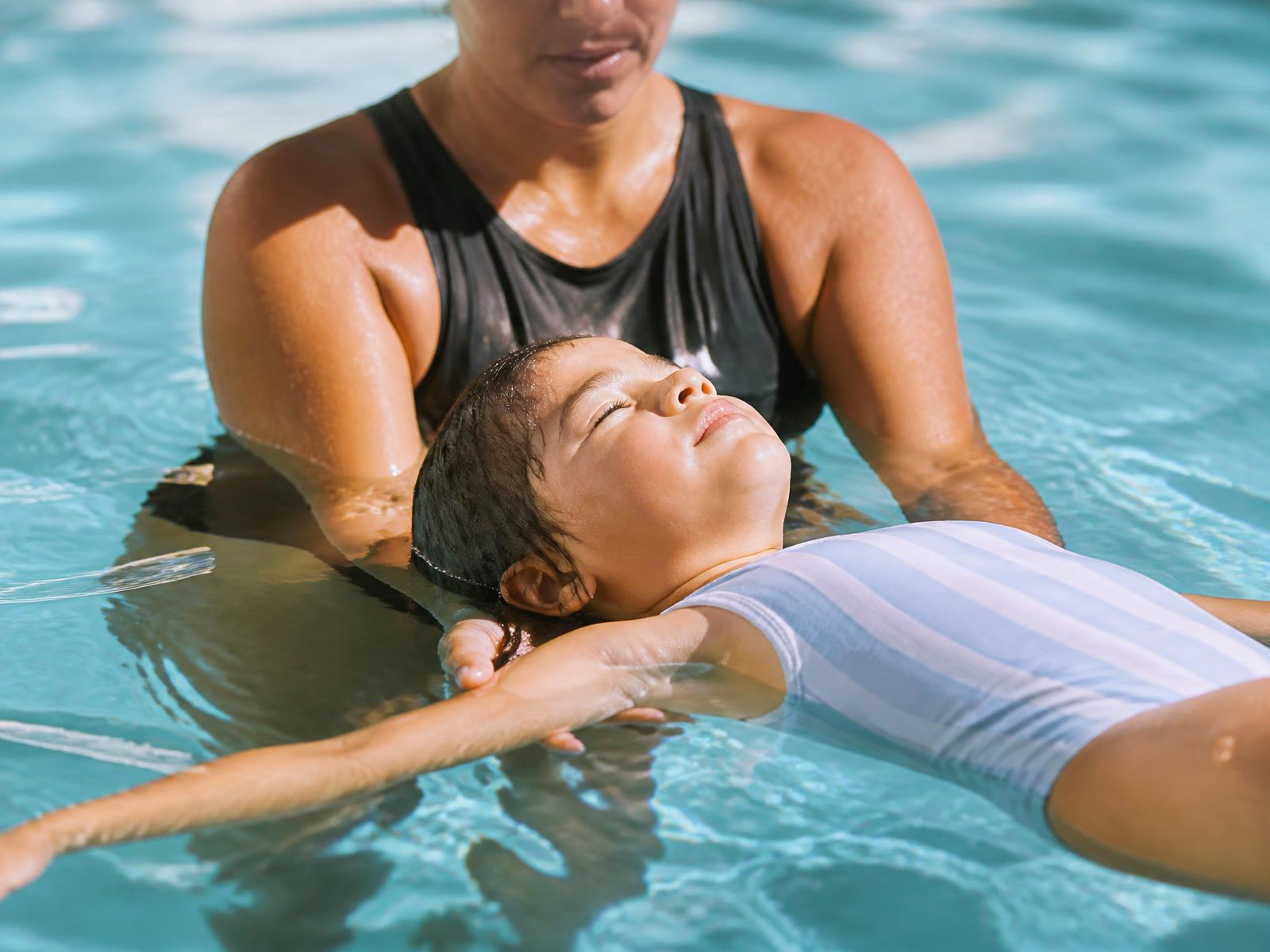 Swim instructor supporting a young child floating calmly on their back in a sunlit pool